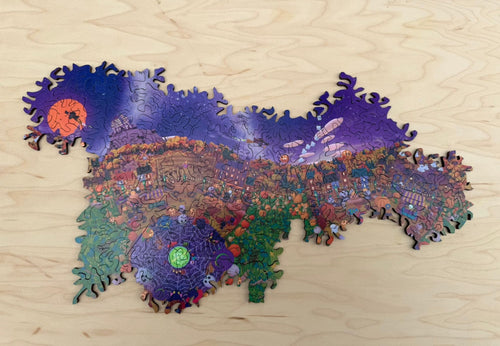 Festival at Shiver Falls infinity puzzle assembled on a wooden table: a Halloween town square with a glowing green cauldron, pumpkin patches, and a purple night sky with an orange moon, edged by curling, borderless pieces